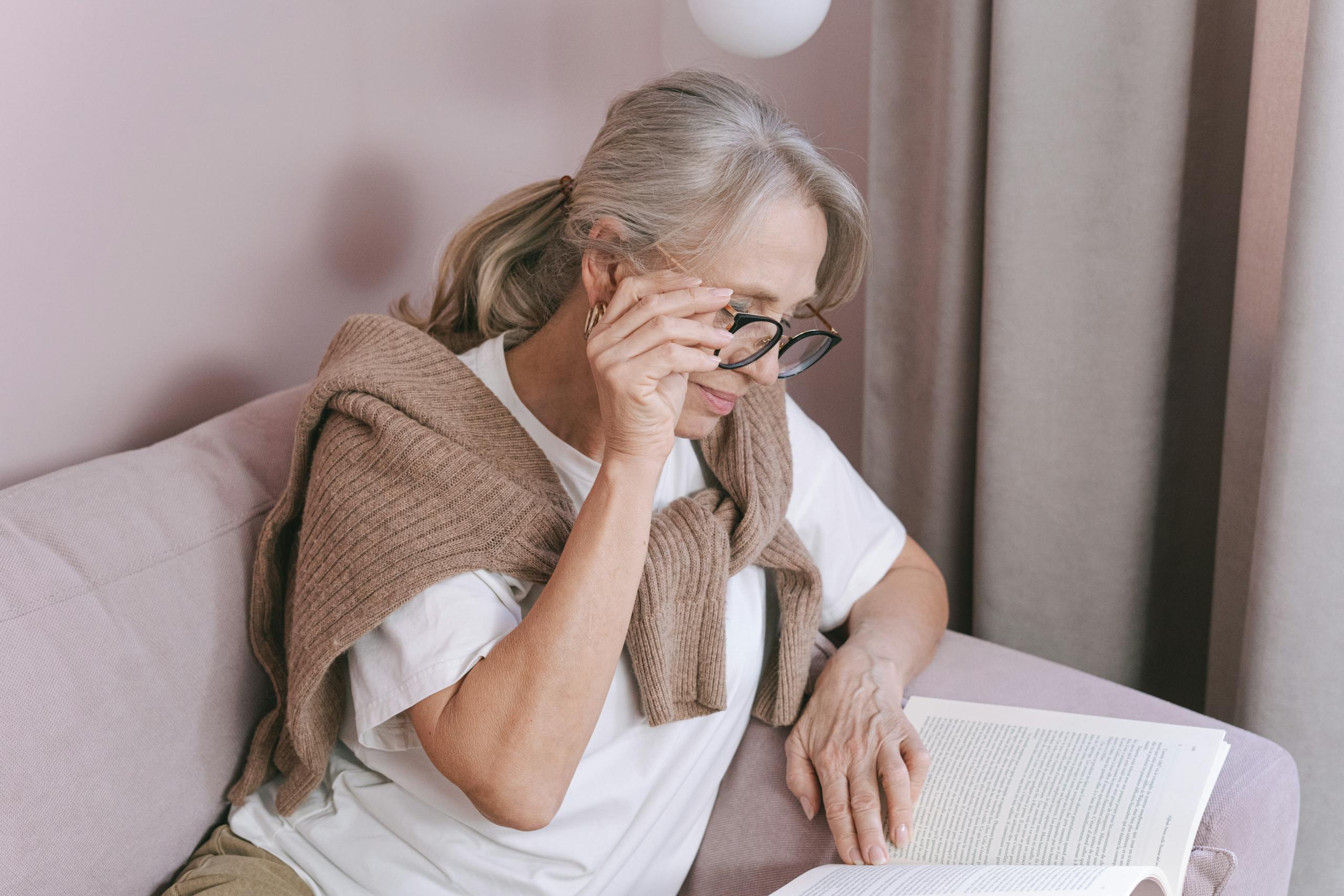 Elderly woman with glasses reading on a couch for leisure at home.