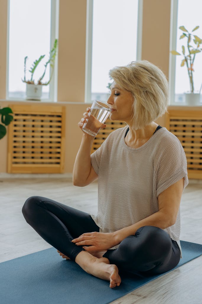 Senior woman in black leggings drinking water while seated on a yoga mat indoors.